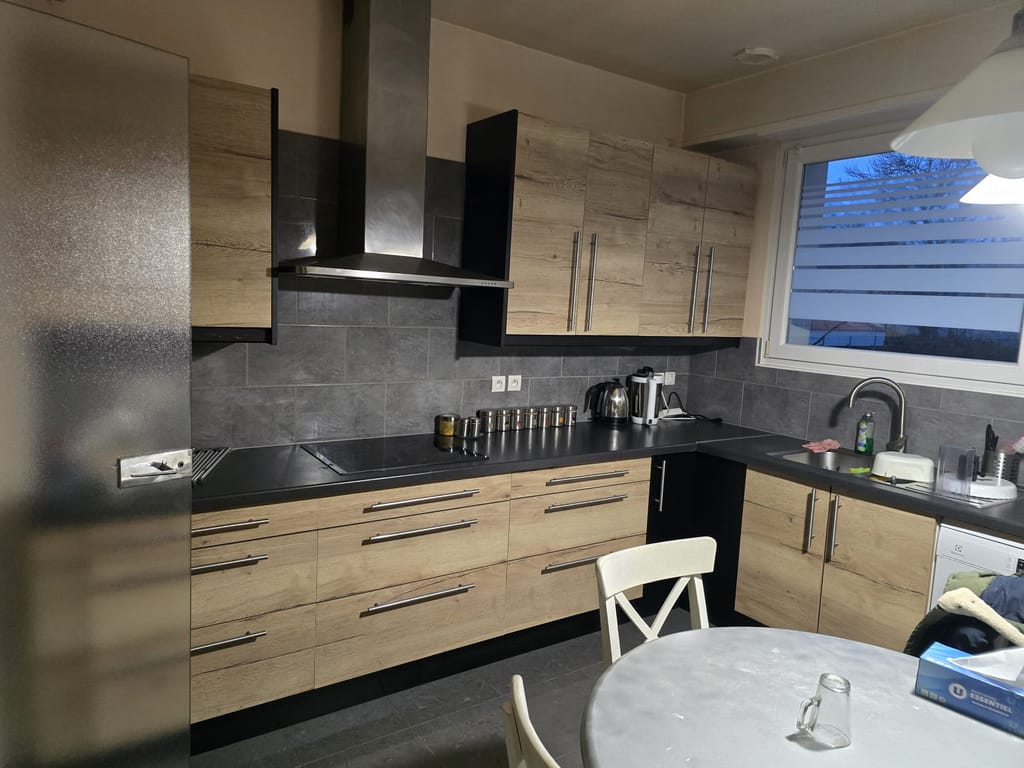 Cuisine en L terminée : façades en chêne clair, plan de travail et colonnes noirs, crédence en carrelage gris et hotte inox au-dessus de la plaque de cuisson.