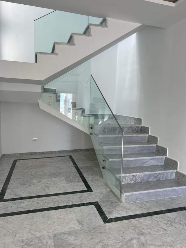 Hall d'entrée terminé : sol en marbre clair encadré par un listel vert foncé, escalier habillé de la même pierre et garde-corps en verre sans montant visible.